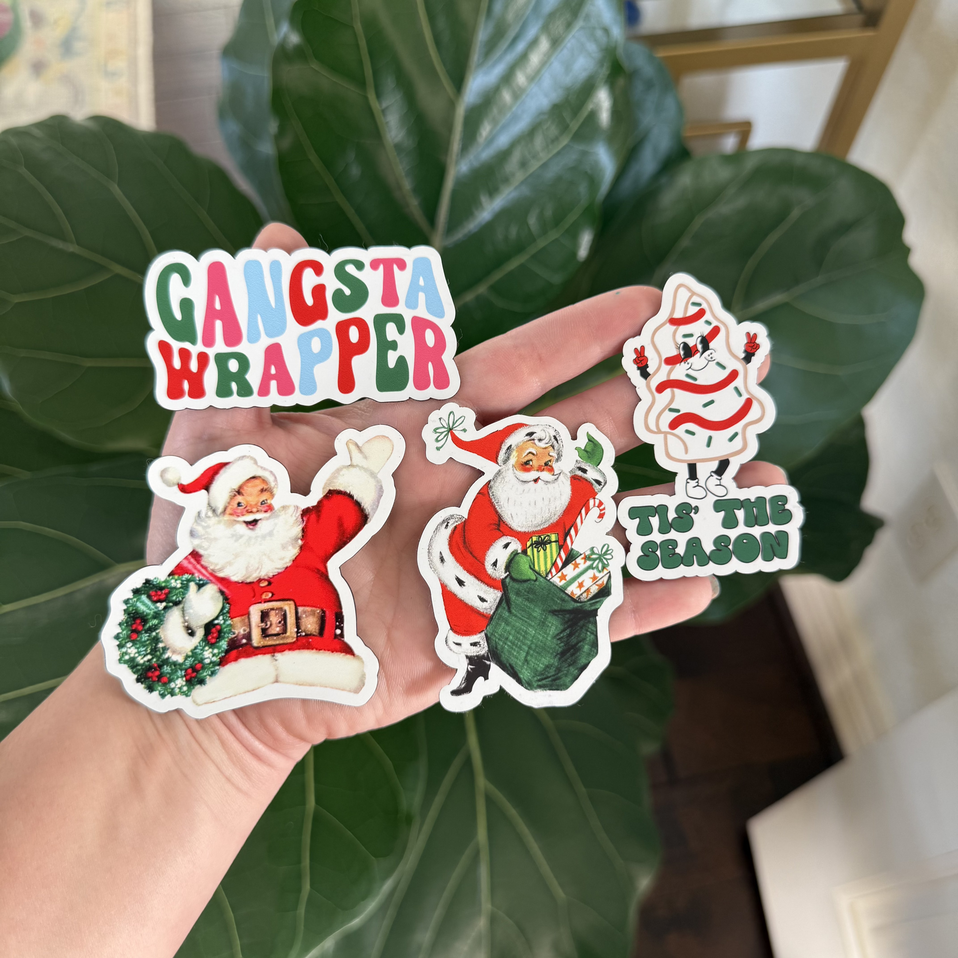 A hand holding four Christmas magnets: one with text reading "Gangsta Wrapper," one with Santa holding a wreath waving, one with Santa and his present sack, and the fourth of a Christmas tree dessert saying "Tis the Season."